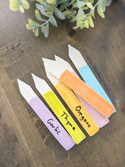 May include: Six pastel-colored ceramic plant markers arranged on a dark brown wooden surface.  Each marker is shaped like a pointed rectangle with a white top and a colored bottom. The colors include light purple, yellow, light orange, and light blue.  Handwritten text on each marker identifies the plant: Garlic, Thyme, Oregano.