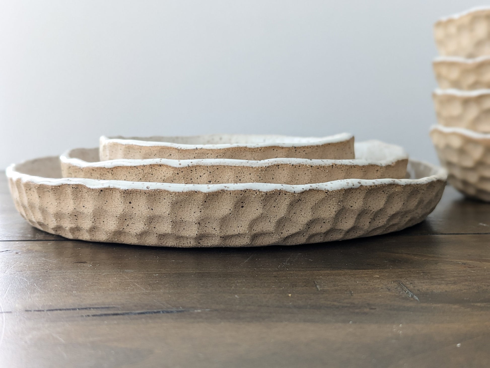 Three nesting ceramic plates of varying sizes are stacked, showcasing a textured, slightly irregular surface with a speckled, off-white rim. The bowls exhibit a rustic, handcrafted aesthetic.